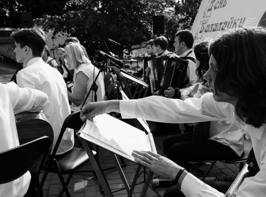 Outdoor orchestral performance with musicians playing various instruments in black and white.