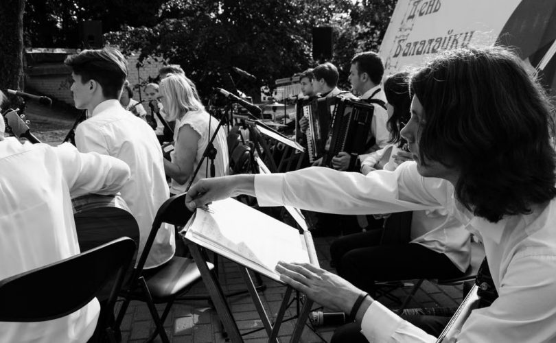 Outdoor orchestral performance with musicians playing various instruments in black and white.